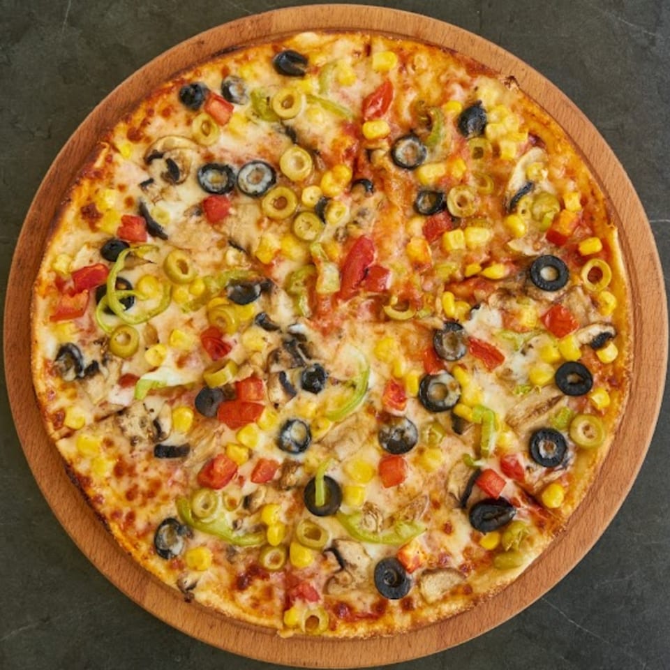 A whole vegetarian pizza with corn, olives, mushrooms, and bell peppers on a wooden board.