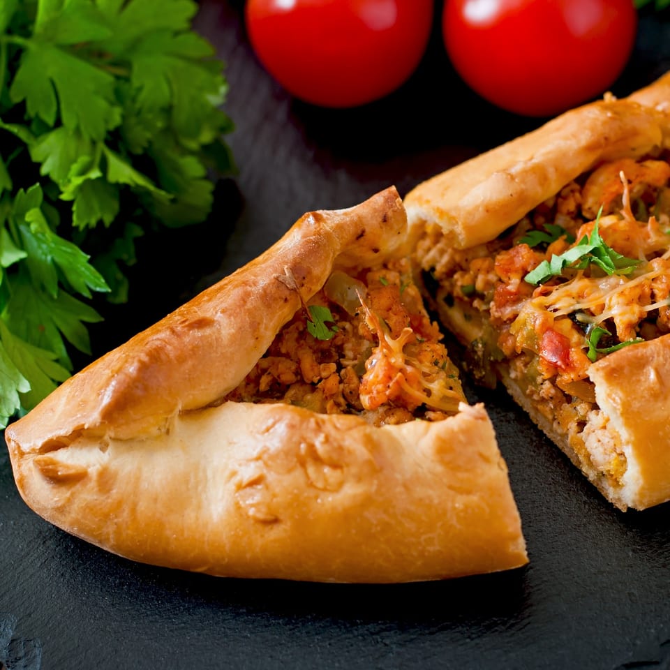 A golden-brown, boat-shaped pizza, cut in half, filled with meat, cheese, and vegetables.