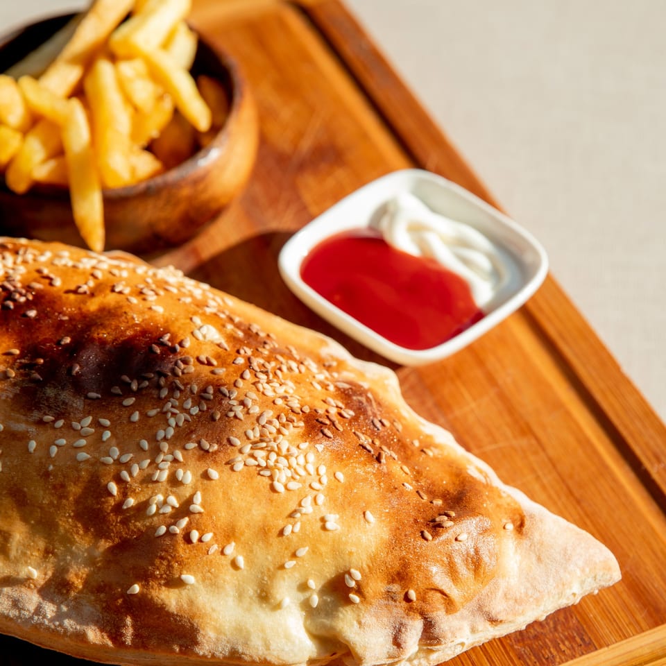 Golden-brown calzone with sesame seeds, a side of fries, ketchup, and mayonnaise on a wooden board.