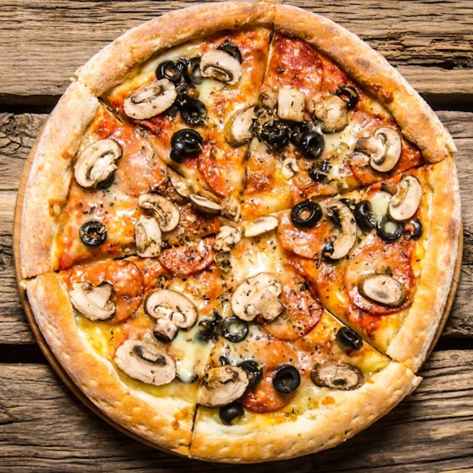 A whole pizza from The PIZZA Shop, topped with mushrooms, black olives, and pepperoni, on a wooden table.