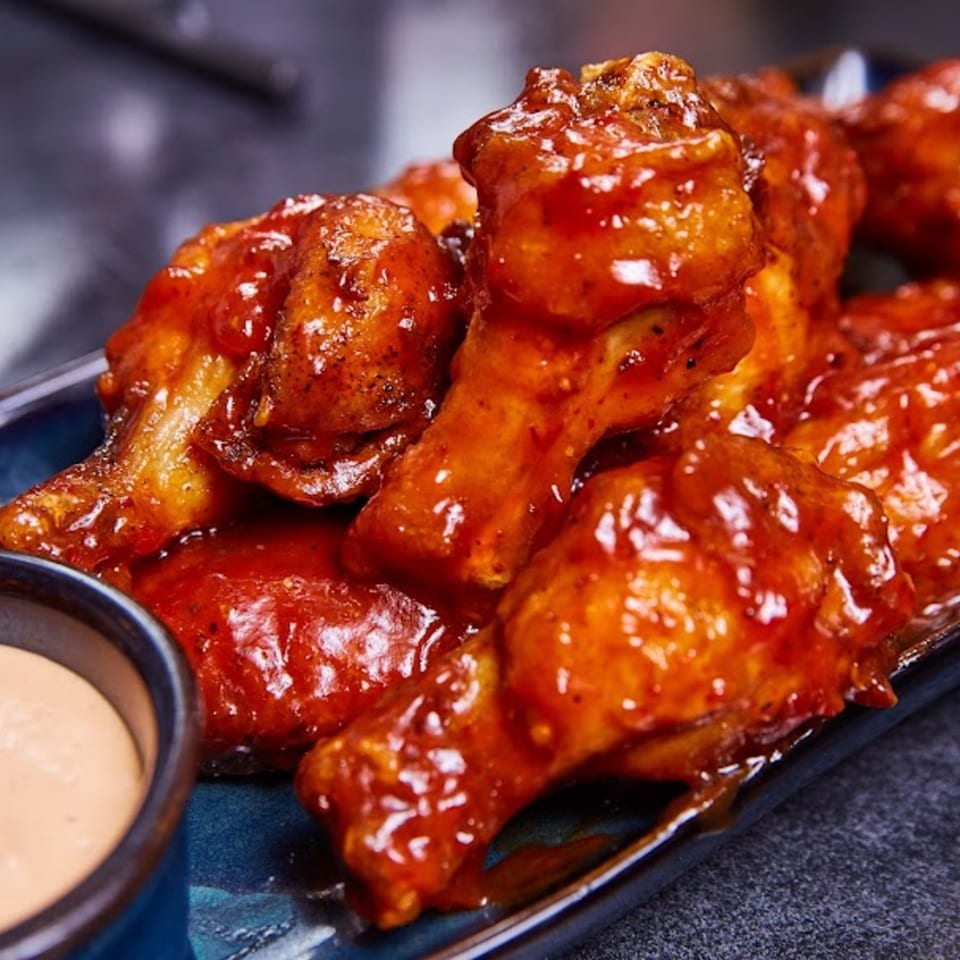 Close-up of several glossy, red-sauced chicken wings on a blue plate with a side of dipping sauce.